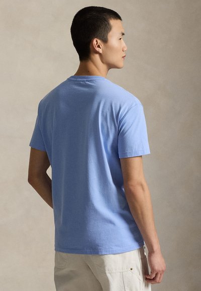 Άντρας που φοράει ένα απλό ανοιχτό μπλε κοντομάνικο μπλουζάκι και μπεζ παντελόνι, στέκεται πλευρικά μπροστά σε ουδέτερο φόντο.