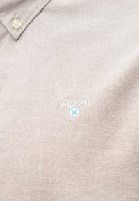 Camisola polo texturada em bege com colarinho abotoado e logótipo "Barbour" bordado em branco acima de um pequeno emblema azul e branco.