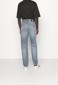 Jeans de mezclilla azul claro con corte recto, bolsillos traseros y un parche negro con el logo en la banda de la cintura. Llevados con zapatillas blancas.