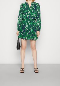 Kukallinen mekko, jossa vihreitä lehtiä tummansiniseen taustaan, pitkät hihat, v-kaula-aukko, vyötärö sidottu, yhdistetty mustiin remmilenkkareihin ja pieneen laukkuun.