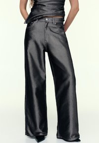 Jean flare en tissu noir métallique, avec une conception classique à cinq poches, taille haute et coutures visibles le long des coutures.