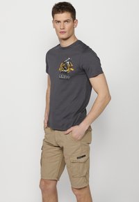 Pilkas medvilninis marškinėlis su grafiniu spaudiniu, derinamas su khaki spalvos šortais iš cargo stiliaus su šoninėmis kišenėmis ir pleistru. Modelis stovi su rankomis kišenėse.