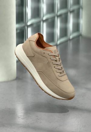 Lichte suède sneaker met beige veters en witte zool, zwevend boven een grijze vloer met een achtergrond van glazen blokken.