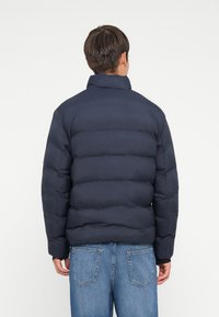 Marineblå puffjakke med quiltet design, høy krage og lange ermer, kombinert med lyseblå denimjeans, sett fra ryggen.