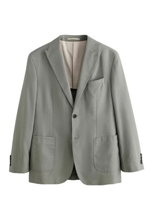 Young man with short dark hair wearing a light gray jacket with a beige collar, facing left with eyes closed against a plain beige background.