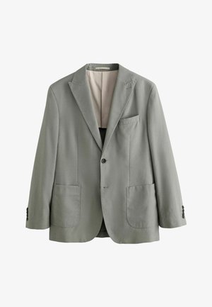 Junger Mann mit kurzen dunklen Haaren, der eine hellgraue Jacke mit beigen Kragen trägt, blickt mit geschlossenen Augen nach links vor einem einfarbigen beigen Hintergrund.