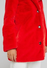 Abrigo de forro polar rojo con una textura suave, que presenta un ajuste entallado, grandes botones negros y bolsillos laterales, el tejido parece mullido y cálido.