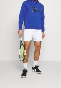 Felpa blu con logo di coccodrillo nero, pantaloni corti bianchi, scarpe da ginnastica nere e calzini bianchi, che tiene in mano una racchetta da tennis con un telaio verde e nero.