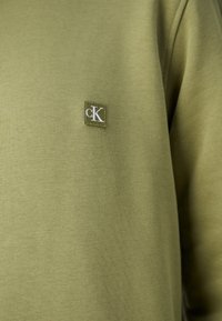 Sudadera verde oliva hecha de tela suave. Presenta un parche cuadrado con el logotipo "cK" en blanco, ubicado en el área del pecho.
