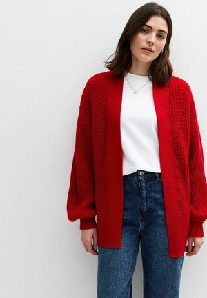 Cárdigan de punto rojo con abertura frontal y mangas oversized, llevado sobre una camisa blanca y jeans de mezclilla azules, mostrado sobre un fondo sencillo.