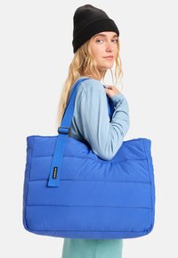 Grote blauwe nylontas met een geborduurd ontwerp, voorzien van een brede schouderriem en een zachte textuur voor comfort.