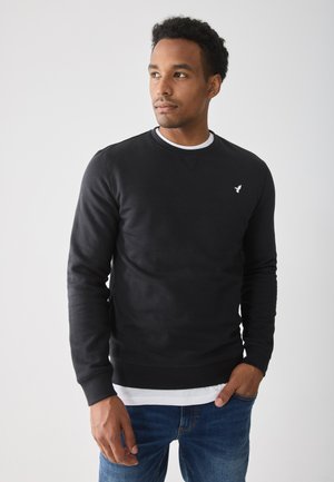 Uomo con un sudario nero sopra una camicia bianca e jeans blu, in piedi con una mano in tasca, che guarda leggermente a sinistra.
