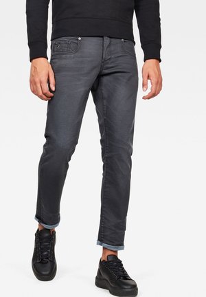 Homme portant un jean gris foncé retroussé, un sweat-shirt noir et des baskets noires, visible de la taille vers le bas sur un fond uni.