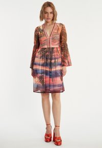 Kurzärmliges, transparantes Kleid mit einem mehrfarbigen Palmenprint, tiefem V-Ausschnitt und Akzentdetails. Getragen mit auffälligen roten Plateauschuhen.