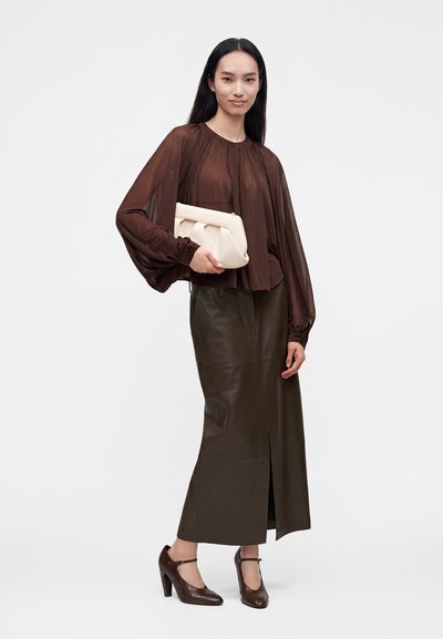 Femme portant un chemisier marron transparent, une jupe midi en cuir sombre avec une fente à l’avant, des talons marron et tenant une pochette blanche.