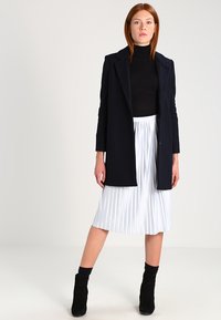 Dunkelblauer Mantel über einem schwarzen Rollkragenpullover, kombiniert mit einem weißen plissierten Midirock und schwarzen Wildleder-Ankle Boots. Einfaches Design, glatte Texturen.