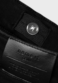 Tela de mezclilla negra con un botón transparente y una etiqueta de cuero negro con texto en relieve. El botón presenta un diseño en forma de cruz.