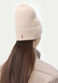 Beige ribbad stickad mössa med vik och en liten etikett; bärs med långt, rakt mörkt hår och en ljus jacka.