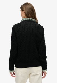 Zwart kabelgebreide trui met geribbelde zoom en manchetten, met daaronder een groen-wit geruit overhemd. Eenvoudig en textuurrijk ontwerp.