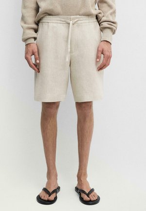 Mann trägt beige Leinen-Shorts mit Kordelzug in der Taille, einen beige gestrickten Pullover und schwarze Flip-Flops an nackten Füßen.