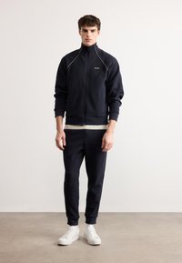 Νεαρός άνδρας στέκεται όρθιος, φοράει μπλε σκούρο τζάκετ με φερμουάρ και ταιριαστό παντελόνι, καθώς και λευκά αθλητικά παπούτσια, μπροστά σε απλό φωτεινό φόντο.