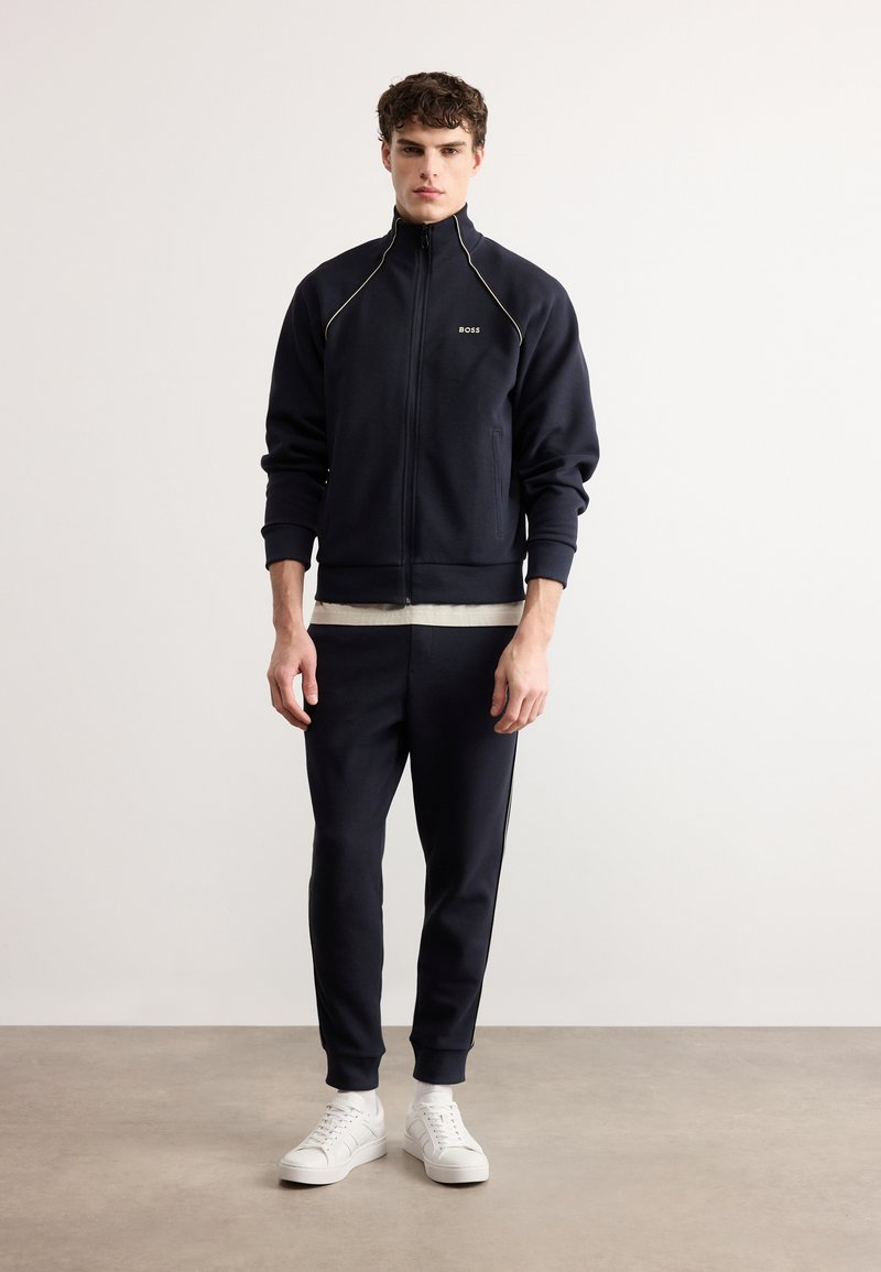 Νεαρός άνδρας στέκεται όρθιος, φοράει μπλε σκούρο τζάκετ με φερμουάρ και ταιριαστό παντελόνι, καθώς και λευκά αθλητικά παπούτσια, μπροστά σε απλό φωτεινό φόντο.