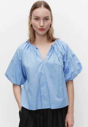 Nainen pukeutuneena vaaleansiniseen liehukehihalliseen V-kauluksiseen paitaan ja mustiin housuihin, seisoo yksivärisen valkoisen taustan edessä.