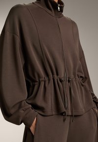 Braune Zip-Jacke mit hohem Kragen, taillierter Passform und Seitentaschen. Das weiche Material verfügt über dezente Paneel-Details und Kordelzüge.