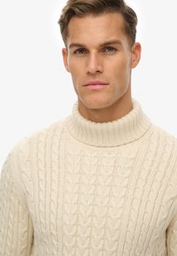 Ung mand med kort brunt hår iført en cremet kabelstrik turtleneck sweater, som ser lige frem med en neutral udtryk.