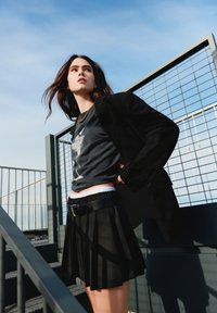 Schwarze Blazer über einem Grafik-T-Shirt, kombiniert mit einem schwarzen, plissierten Rock, der einen strukturierten Bund und einen Gürtel hat. Blauer Himmel im Hintergrund.