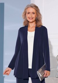 Dunkelblauer langer Cardigan mit offener Vorderseite und langen Ärmeln, über einem weißen Oberteil getragen. Hält eine silberne Clutch. Glatte Textur, keine Muster.