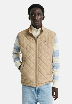 Gilet matelassé beige avec un col montant, fermeture éclair sur toute la longueur, porté sur un pull en tricot à gros torsades multicolore. Dispose de coutures diagonales et d'un logo.
