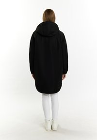 Giacca nera con cappuccio dal design lungo e dritto; presenta polsini elasticizzati e orlo arrotondato, indossata sopra pantaloni bianchi e sneakers.