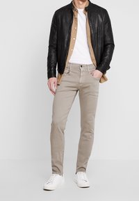 Chaqueta de cuero negra, camisa de botones color marrón claro, camiseta de cuello redondo blanca, pantalones beige claros y zapatillas blancas. Diseño simple y de corte ajustado.