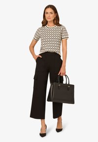 Schwarze weite Cargohosen, gemustertes cremfarbenes Oberteil und eine strukturierte schwarze Handtasche mit klarer Linienführung und goldenen Akzenten.