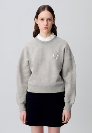 Sudadera gris con un cuello y puños acanalados, con un pequeño logo bordado. Llevada sobre una blusa blanca con volantes y una falda negra.
