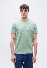 Νέος άνδρας που φοράει ανοιχτοπράσινο κοντομάνικο T-shirt με το λογότυπο "BOSS" στο στήθος και μπλε τζιν, στέκεται μπροστά σε λευκό φόντο.