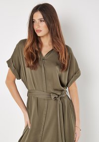 Olivegrünes, mit Gürtel gebundenes Kleid aus weichem, leichtem Stoff. Verfügt über kurze umgeschlagene Ärmel und eine knöpfbare Vorderseite mit vertikalen Nähten.