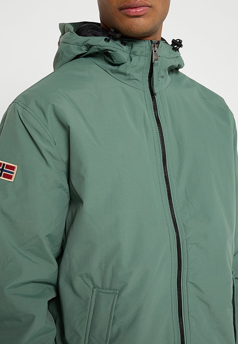 Veste imperméable verte avec fermeture éclair, capuche et doublure noire contrastante. Elle présente un patch de drapeau brodé sur la manche gauche.