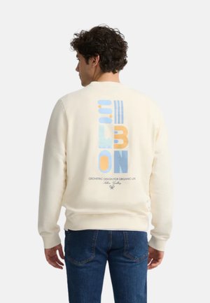 Sudadera crema con un gráfico geométrico en azul y naranja en la parte trasera, con puños y dobladillo acanalados. Combinada con jeans azules.