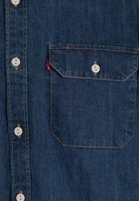 Tummansinisestä denim-paitasta, jossa on terävä kaulus, viisi valkoista nappia ja läpäntasku. Huomaa kontrastoiva ommel ja sivuetiketti.