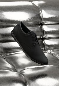 Zapato casual negro con cordones descansando sobre una superficie metálica brillante con cojines plateados reflectantes al fondo.