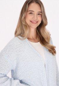 Cardigan de color azul claro, suave, con un diseño de escote en V y frente abierto. Tejido de punto texturizado, corte relajado y hombros caídos.