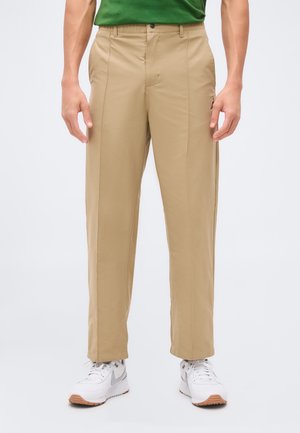 Man draagt beige rechtvallende broek, witte sneakers en een groen overhemd, handen ontspannen langs het lichaam tegen een effen achtergrond.