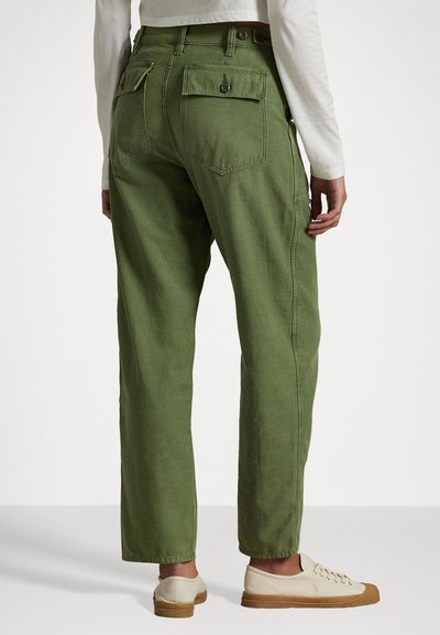 Pantaloni di cotone verde con una vestibilità rilassata, dotati di due tasche posteriori e dettagli di cucitura laterale. Indossati con scarpe chiare.