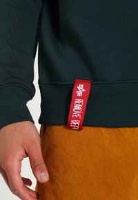 Sudadera de color verde oscuro con puños acanalados, que presenta una etiqueta de tela roja prominente con la inscripción “REMOTE BEEF” en el dobladillo. Material de mezcla de algodón.