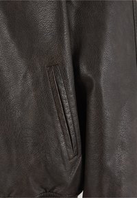 Jachetă din piele maro cu finisaj texturat, cu buzunar vizibil și tiv striat. Pielea pare moale și flexibilă.