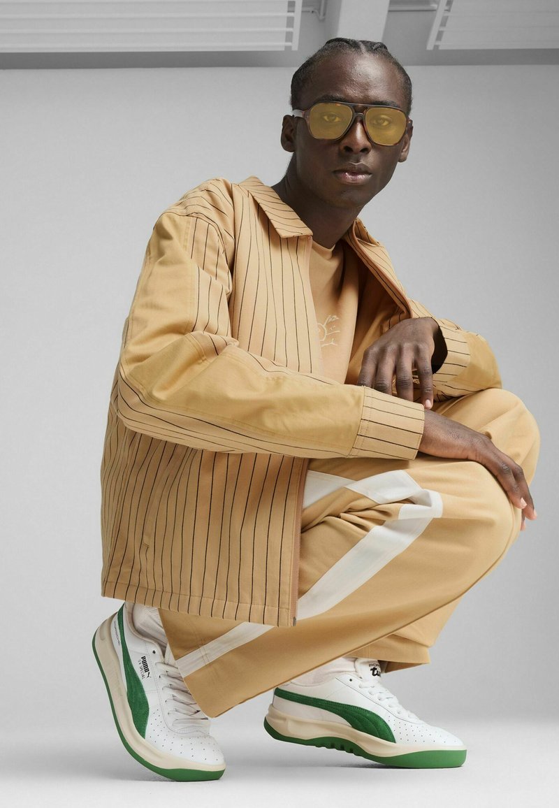 Veste rayée beige sur une chemise beige, associée à un pantalon beige avec des rayures blanches sur les côtés. Baskets blanches avec des accents verts. Lunettes de soleil jaunes.