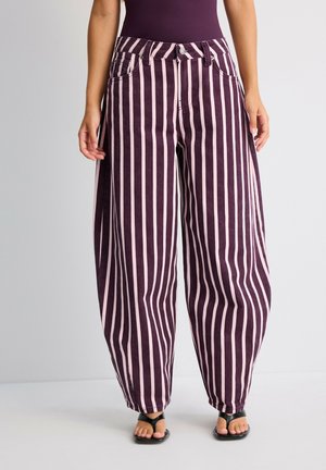 Femme portant un pantalon ample à rayures verticales roses et violet foncé avec des sandales noires, debout devant un fond uni.