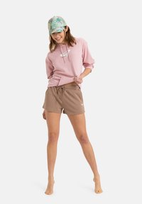 Hoodie rose avec cordons et logo, associé à un short marron. Le mannequin porte une casquette à fleurs vertes. Chaussures : pieds nus.